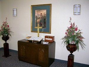 Funeral home interior. Dark wooden cabinet with open book and gold cross. Floral arrangements flank it, painting above.