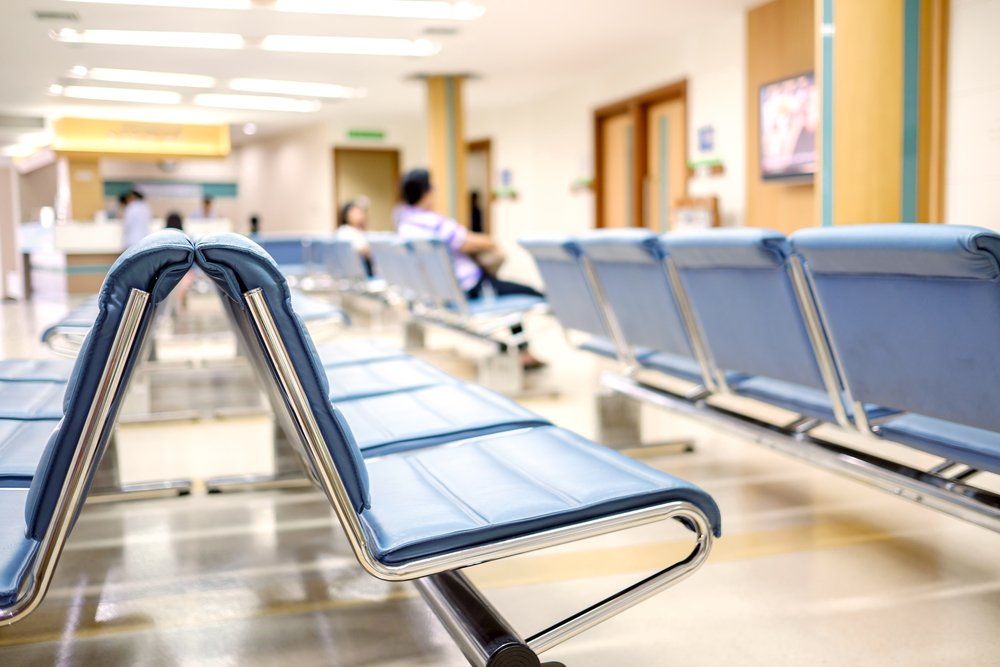 Empty blue chairs in a brightly lit waiting room; a few people sit further back.