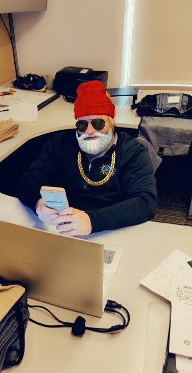 A man with a fake beard is sitting at a desk looking at his phone.