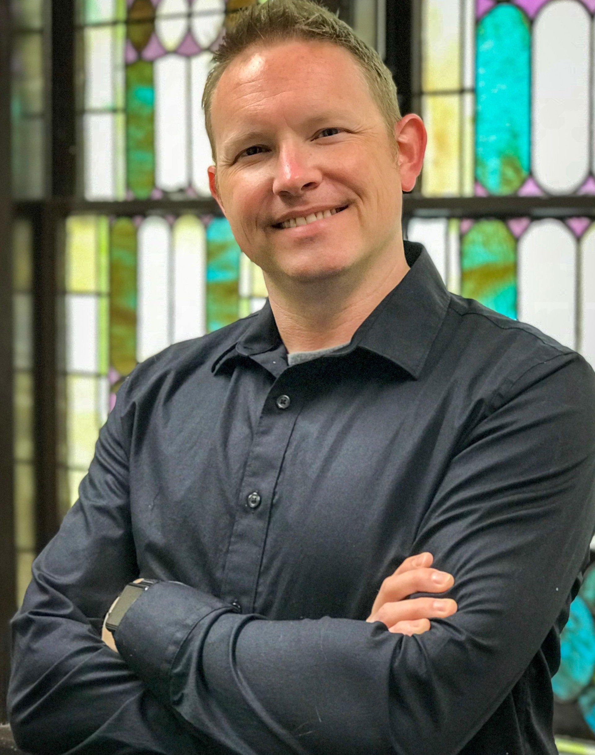 A man in a black shirt is standing with his arms crossed in front of a stained glass window.