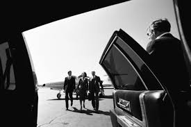 People in suits exiting a car onto an airfield. A private jet is visible in the background.