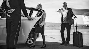 Man opens car door for woman near a private jet; man with luggage stands nearby.
