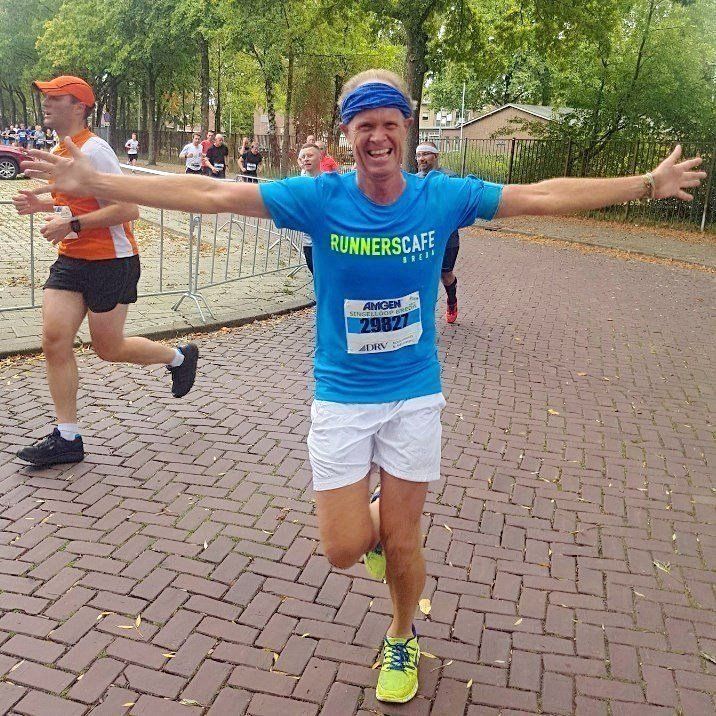 Een hardloper in een blauw shirt en witte korte broek, met uitgestrekte armen, viert de finish op het bakstenen pad.