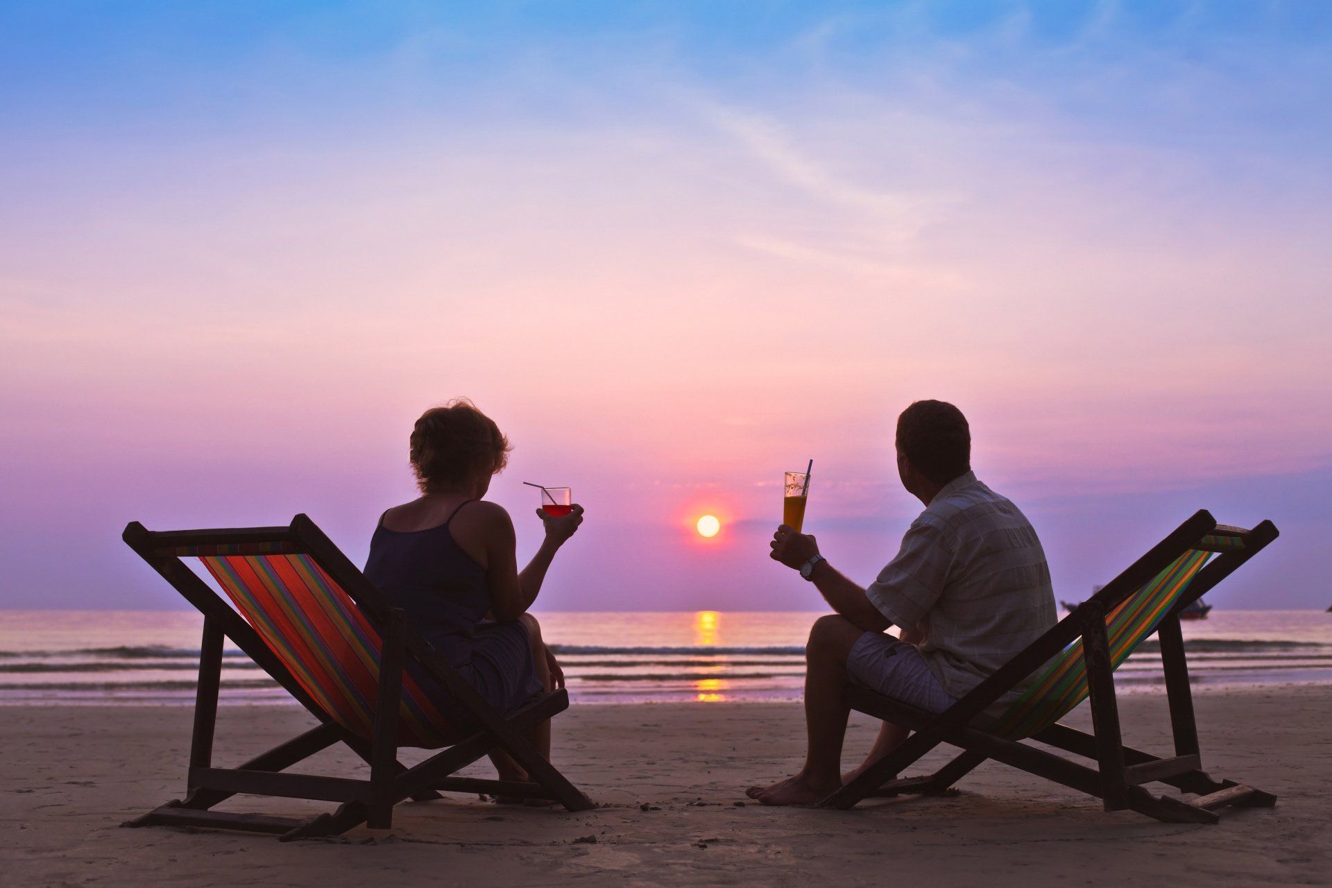 A man and a woman are sitting in beach chairs on the beach drinking cocktails.