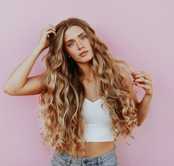 A woman with very long hair is standing in front of a pink wall.