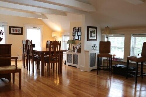 A bright, wood-floored dining and living space with a wooden table, chairs, and lamps under vaulted ceilings.