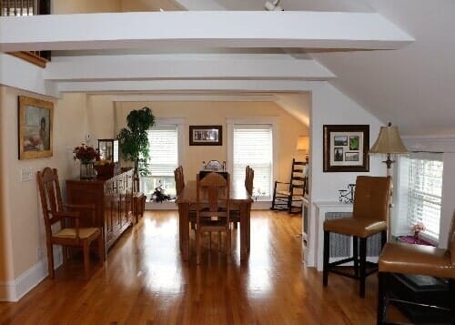 A warm, brightly lit dining area with hardwood floors, a wooden table, chairs, and large windows under a vaulted ceiling.