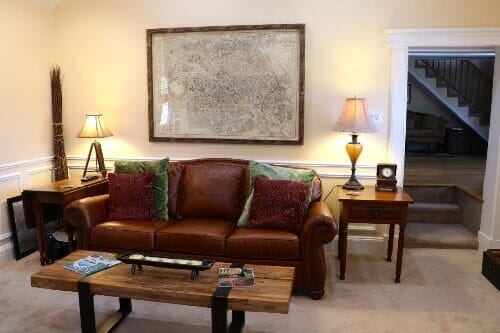 A brown leather couch with patterned pillows sits in a living room with a rustic coffee table, lamps, and a wall map.