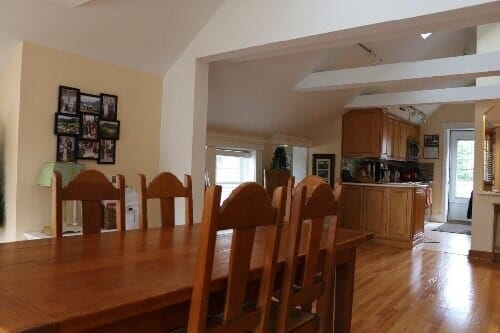 A wooden dining table and chairs in the foreground, with an open-plan kitchen and a hallway visible in the background.