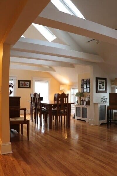 A dining room with a wooden table and chairs, hardwood floors, exposed ceiling beams, and multiple skylights.