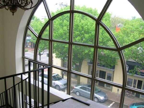 A large, arched window overlooking a tree-lined city street with parked cars, viewed from a high indoor stair landing.