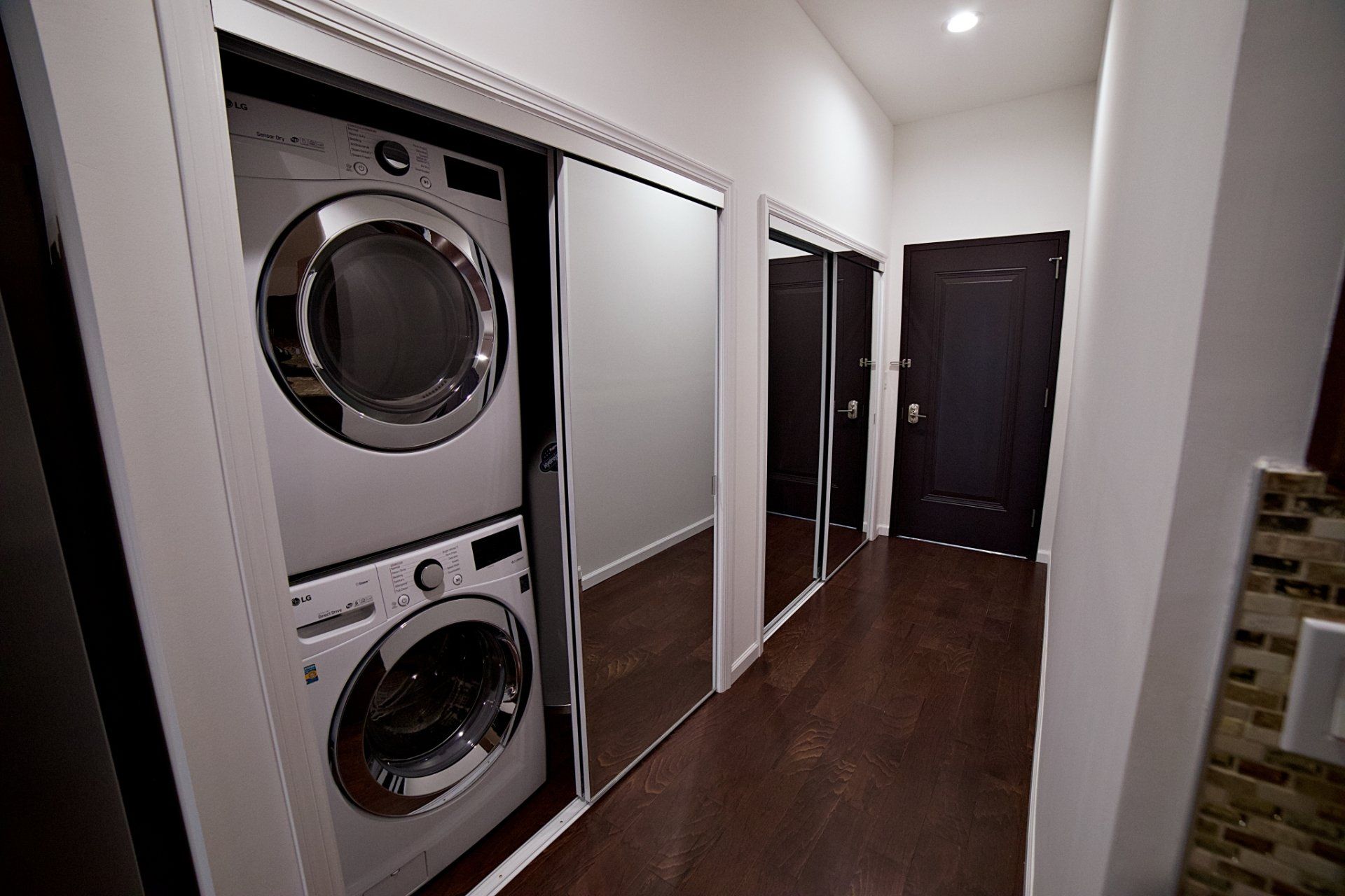 A stacked white washer and dryer in a hallway closet next to mirrored sliding doors, leading to a dark brown door.
