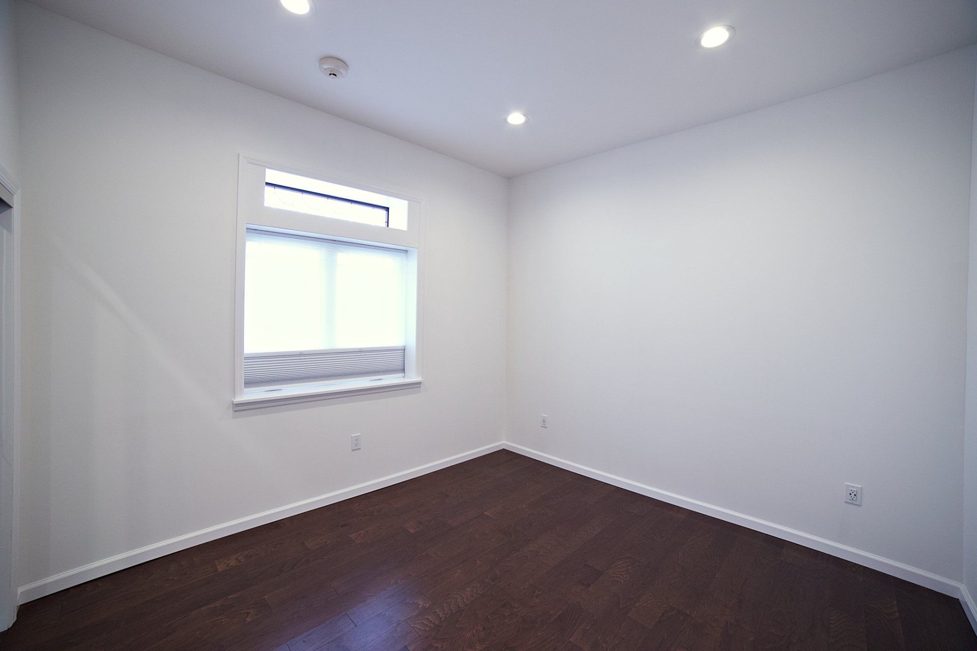 An empty room with white walls, dark wood floors, recessed ceiling lights, and a window with a blind.