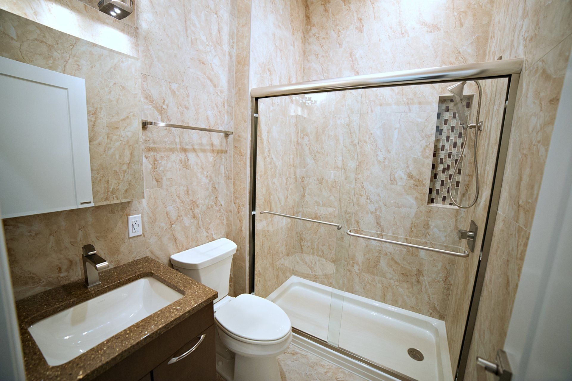 A modern bathroom featuring beige marble-patterned walls, a white vanity sink, a toilet, and a glass-enclosed shower.
