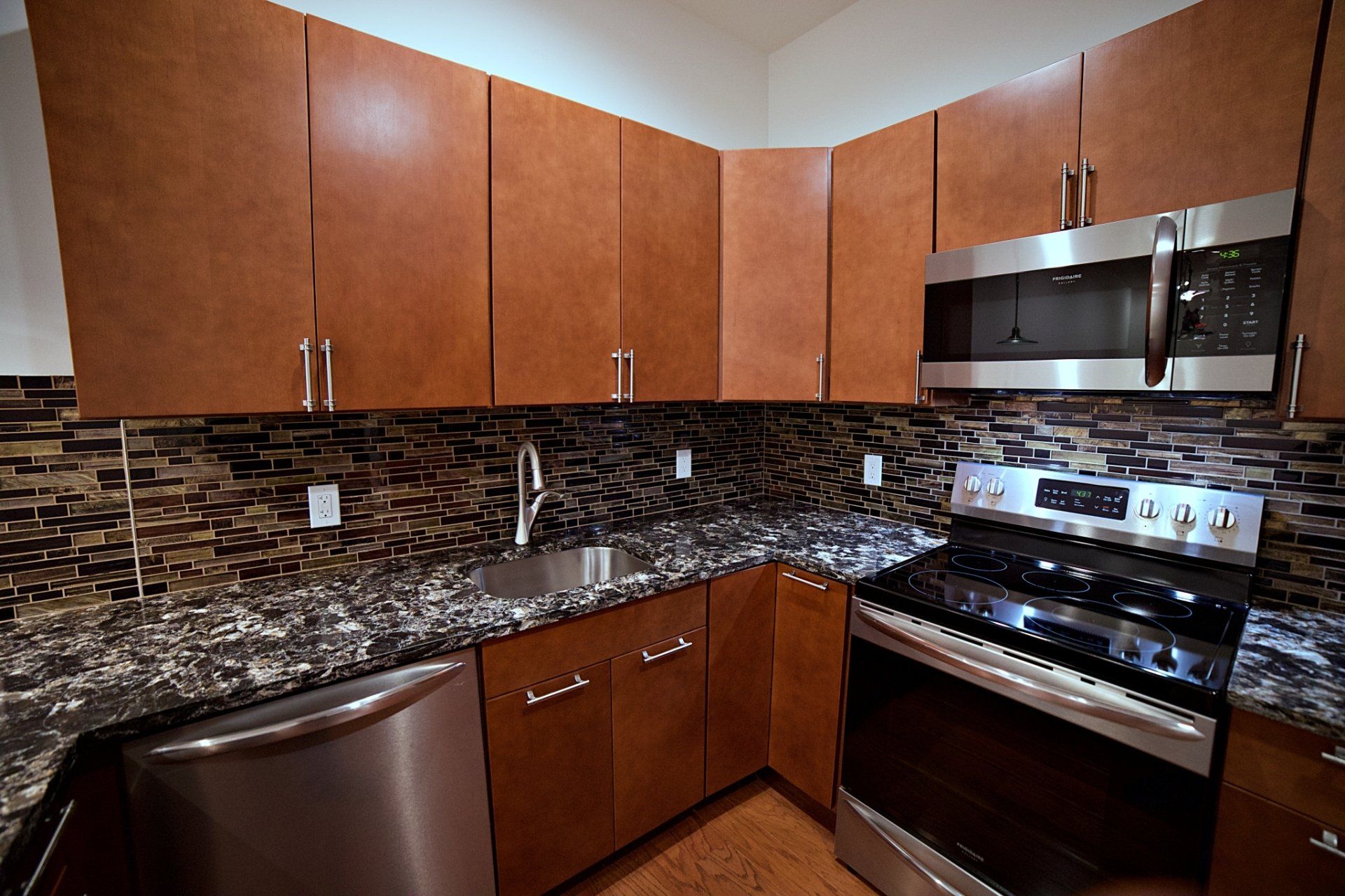 Modern kitchen with brown cabinets, stainless steel appliances, a stone tile backsplash, and dark granite countertops.