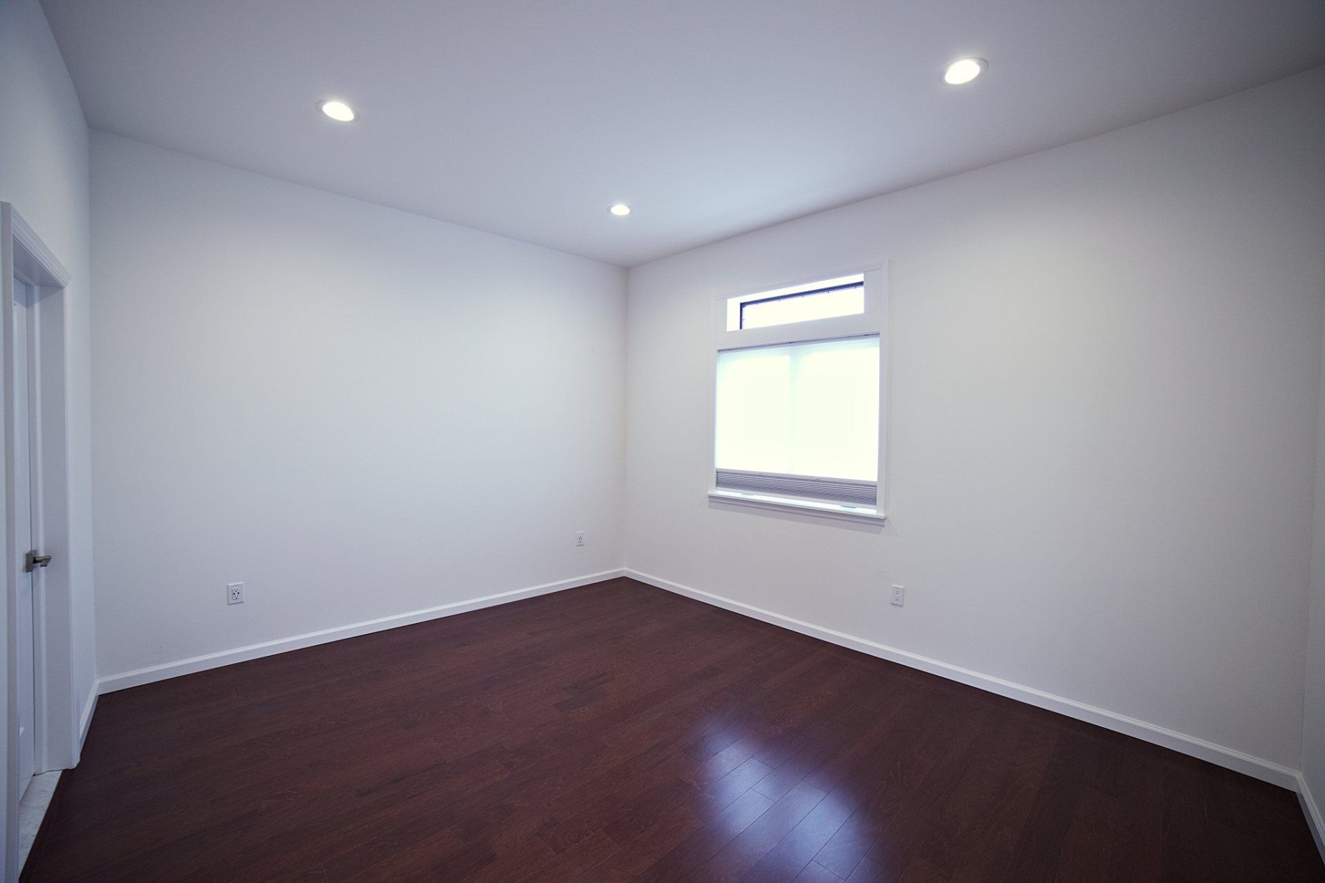 An empty bedroom with white walls, recessed ceiling lights, dark hardwood floors, and a window with a blind.