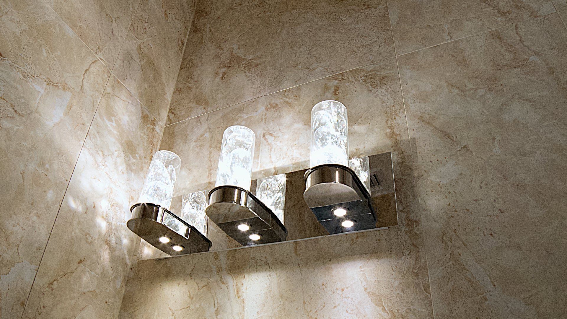 A three-light vanity wall fixture with textured glass cylinders and a metallic base, mounted on a tan stone tile wall.