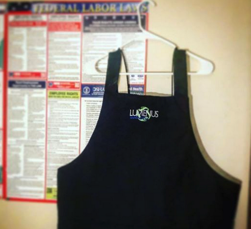 A black apron with the word luvenus embroidered on it hangs on a hanger