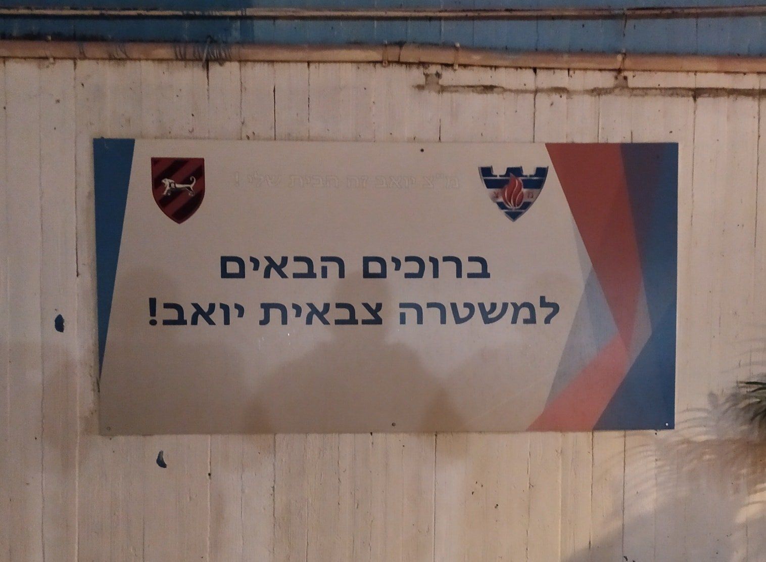 שלט עם טקסט וסמלים בעברית על רקע לבן, המברך את המשטרה הצבאית, עם נגיעות כחולות ואדומות.