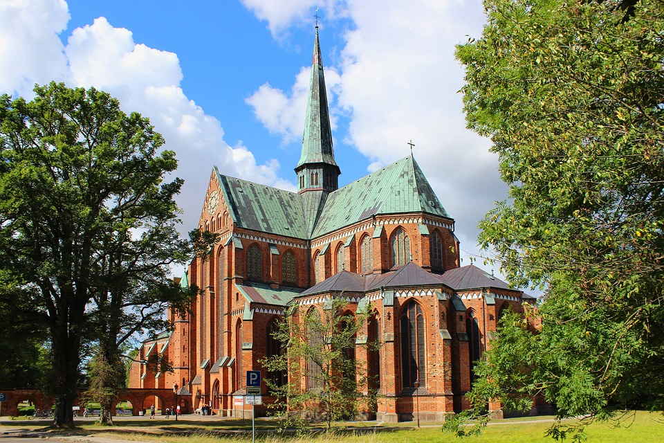 pixabay, lizenzfrei Eine große Kirche aus rotem Backstein mit grünem Dach ist von Bäumen umgeben.
