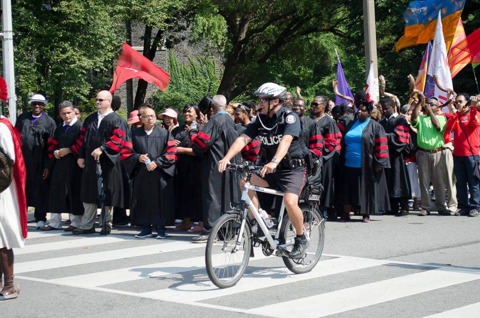 man in a cycling outfit, cycling past crowds at a parade, the crowd has many different colour flags