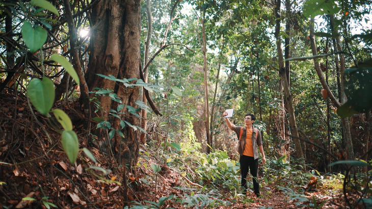 Person in orange jacket standing on a forest trail, surrounded by dense green trees and foliage