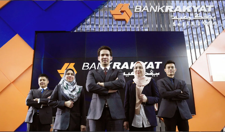 Five people posing onstage in front of a Bank Rakyat backdrop, arms crossed, in formal attire