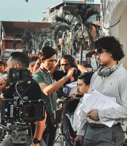 Film crew filming outdoors with camera rig, palm trees, and a person holding papers on the right