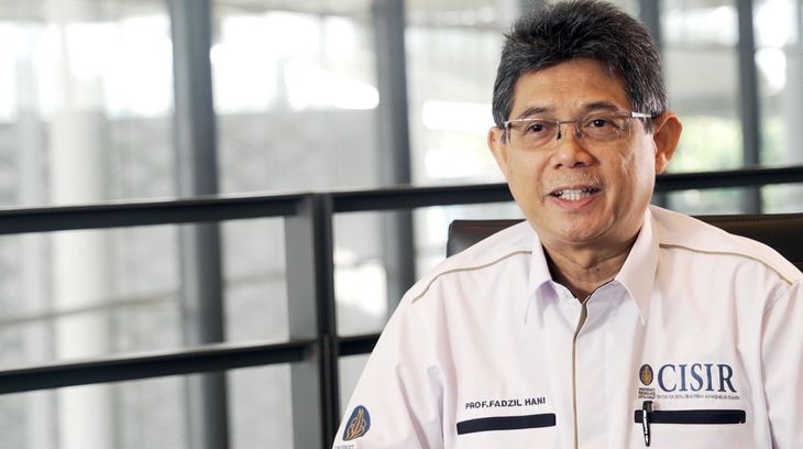 Man in a white collared shirt with an OCISIR logo, standing indoors near a railing and windows.