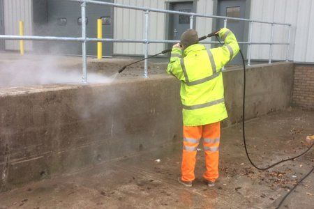 A man in a yellow jacket and orange pants is cleaning a wall with a high pressure washer.