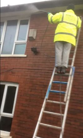 A man in a yellow jacket is standing on a ladder in front of a brick building.