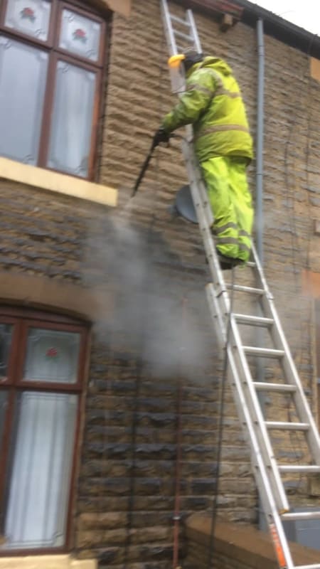 A man is standing on a ladder cleaning a brick building with a high pressure washer.