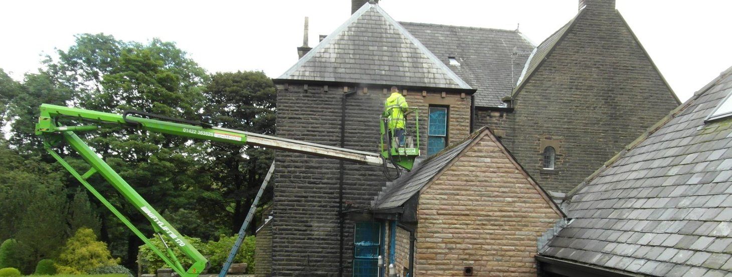A man is standing on a green crane in front of a brick building.