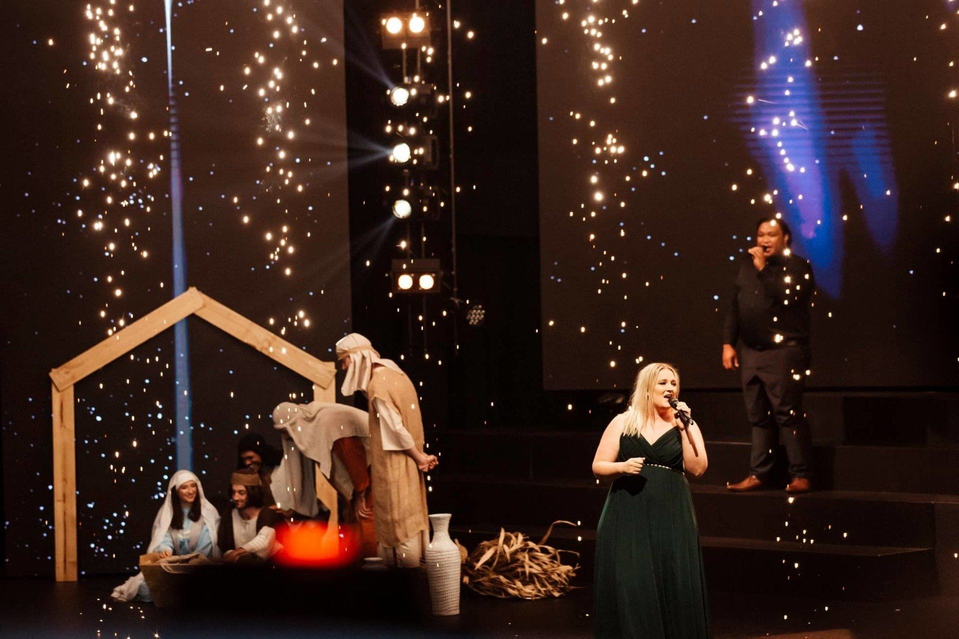 A woman in a green dress is singing on a stage in front of a nativity scene.