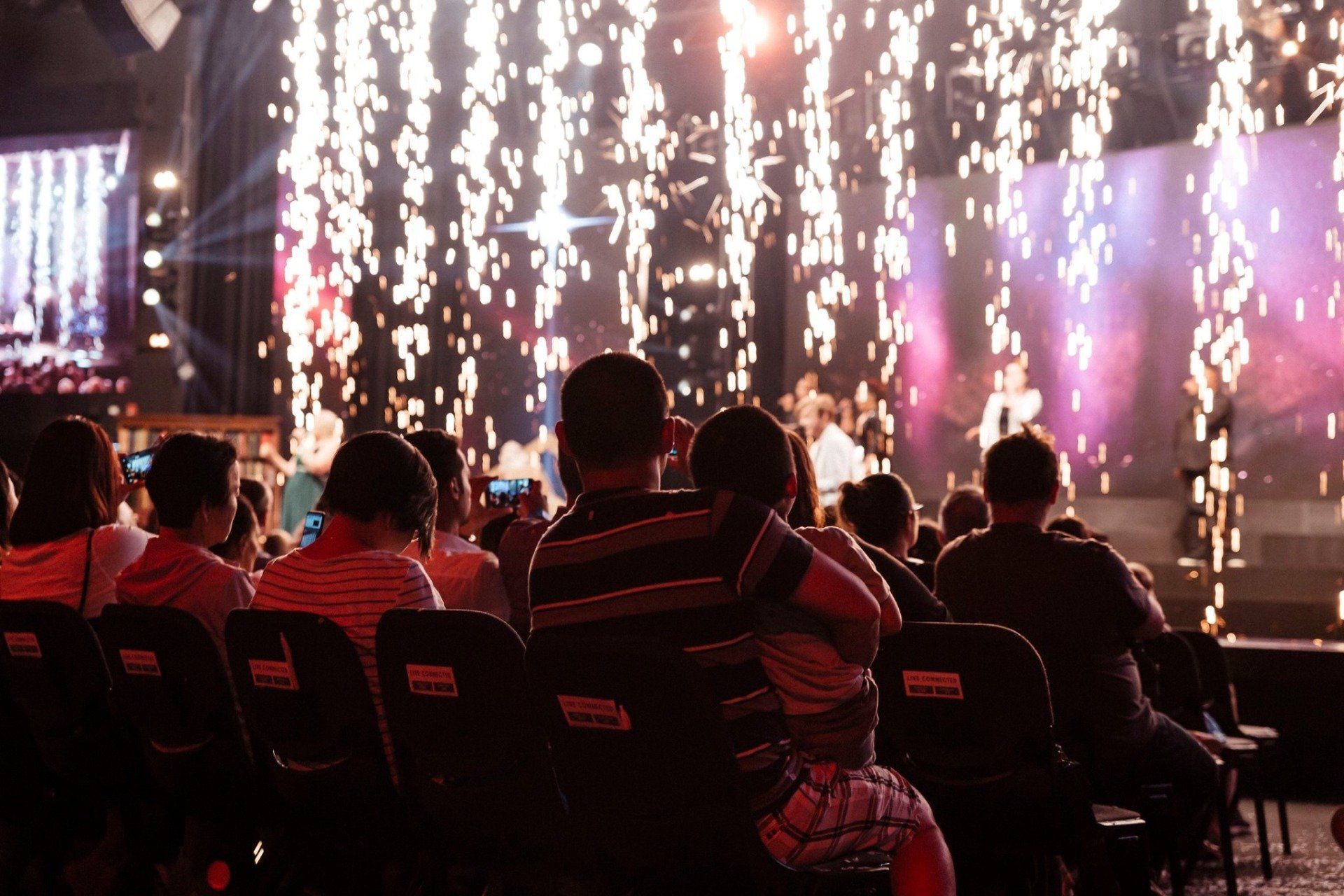A group of people are sitting in a stadium watching a concert. with stage Pyrotechnics 