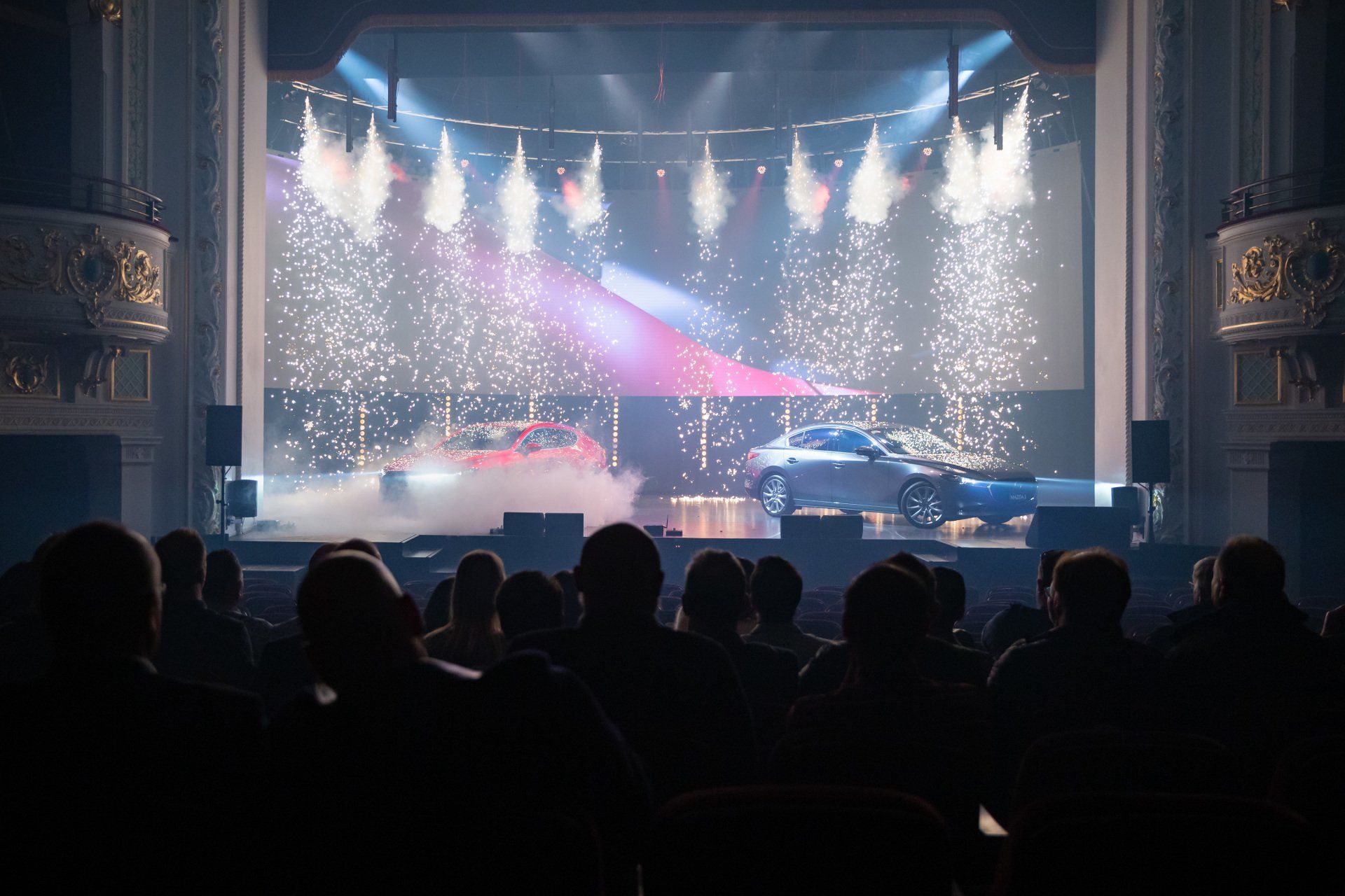 A group of people are sitting in front of a stage with a car on it with pyrotechnics