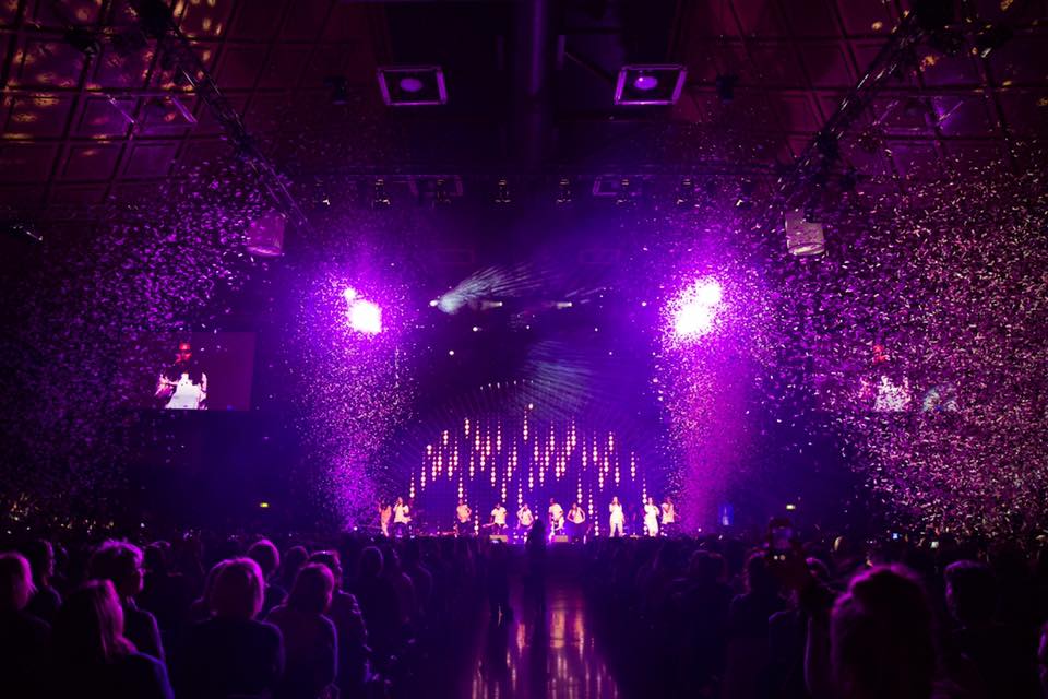 A crowd of people are watching a concert in a stadium with purple lights.