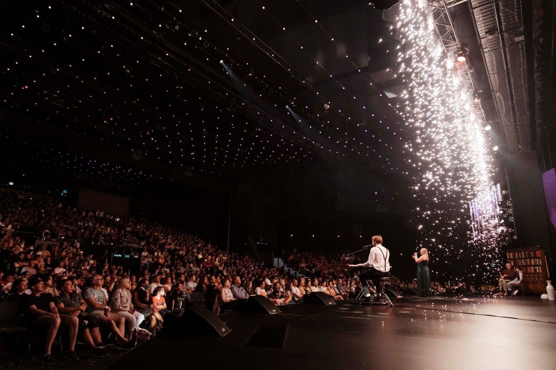 A man is standing on a stage in front of a crowd of people watching a fireworks display. Stage pyrotechnics 