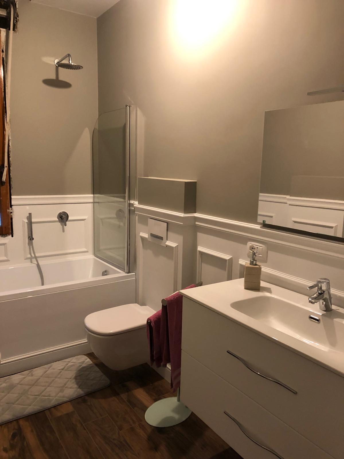 Bagno con vasca bianca, WC e lavandino. Pareti grigie, pavimento effetto legno e soffione doccia.