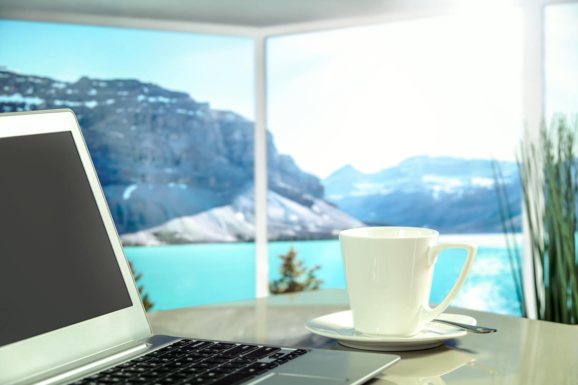 A laptop and a cup of coffee on a table in front of a window.