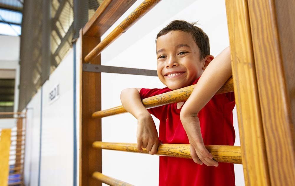 A young boy is leaning on a wooden bar in a gym and smiling.