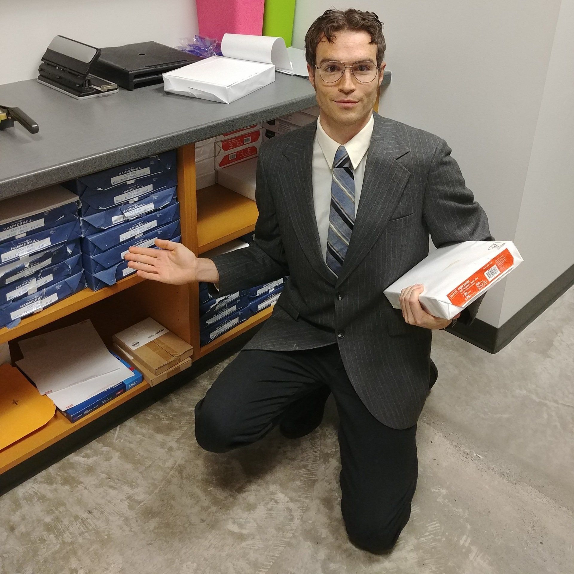 Man in suit kneels near a shelf of paper, holding a ream and gesturing with his other hand.