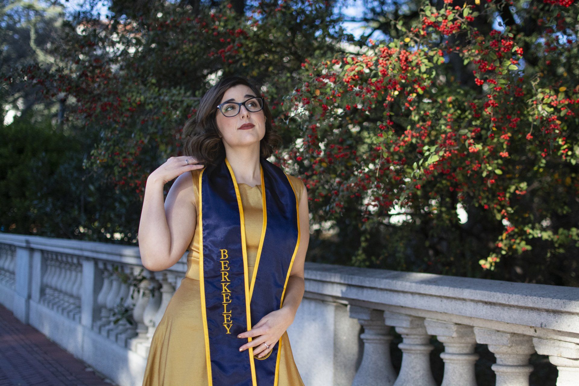 Woman in glasses and a gold dress with a blue sash stands on a stone balcony, looking up. Red berries and greenery in the background.
