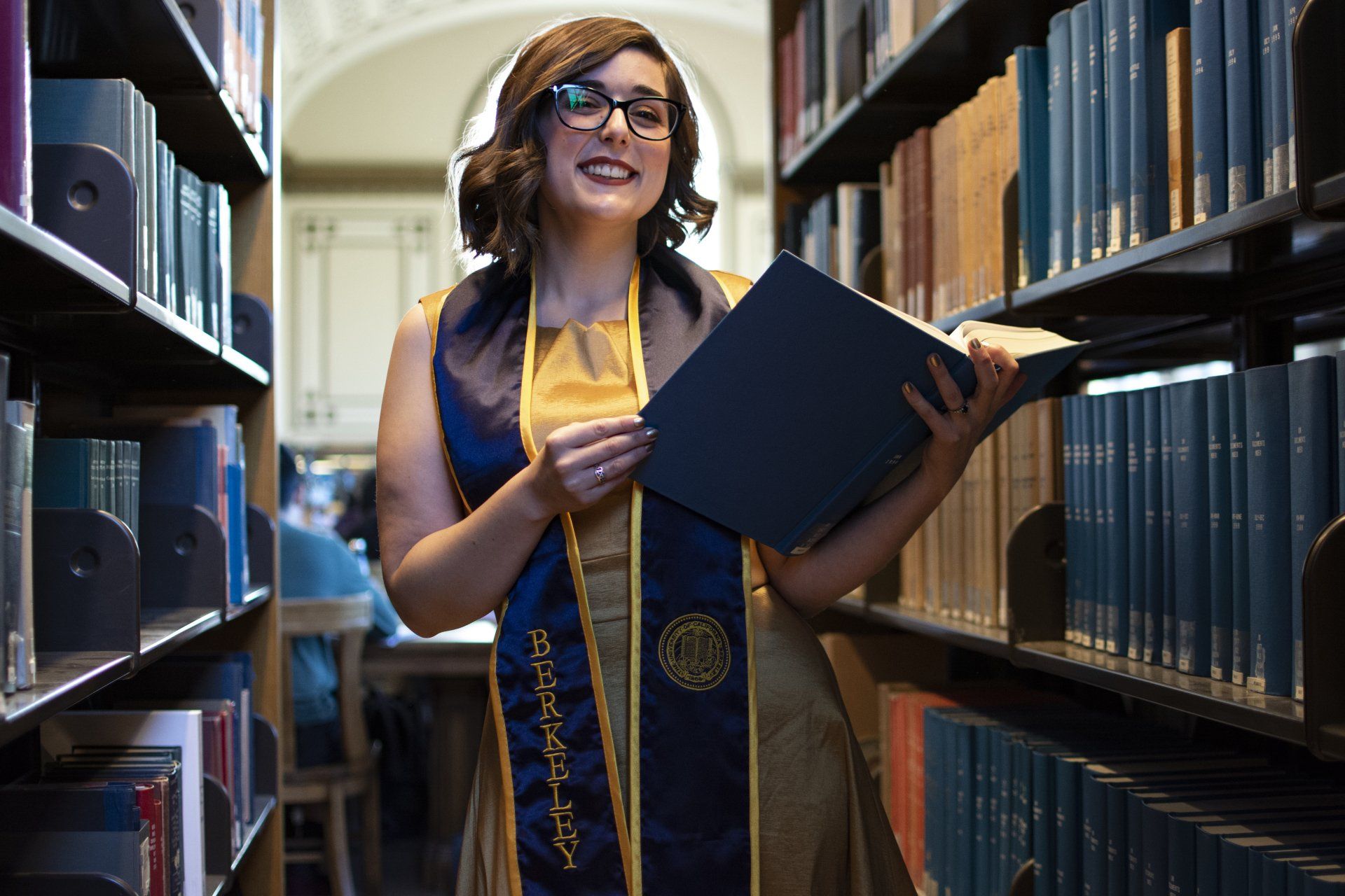 Woman in gold gown and sash, holding a book, smiles in a library aisle.