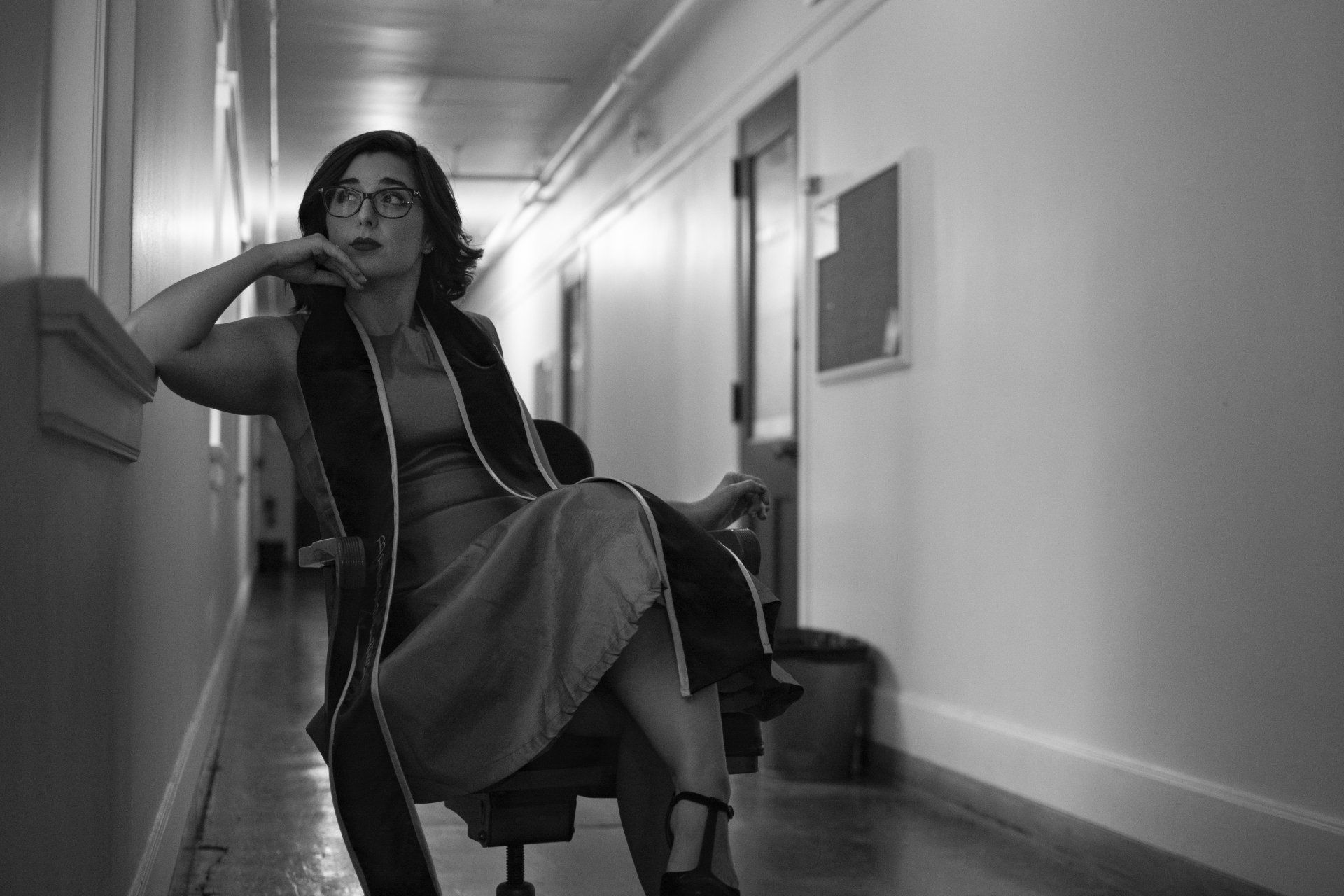 Woman in glasses sits in a chair in a hallway, looking thoughtful. Black and white.