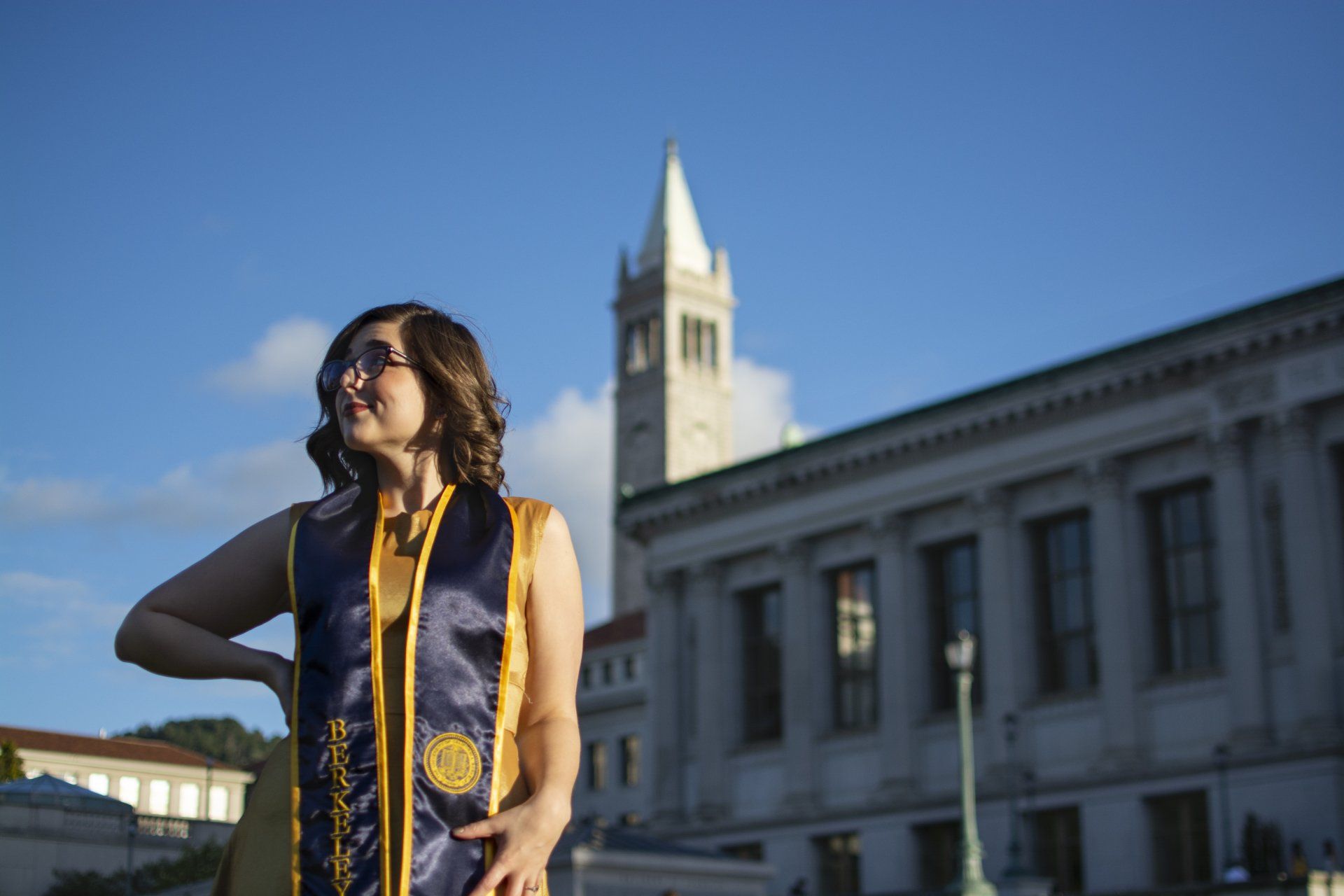 Woman in graduation gown looking upward at a campus building under a blue sky.