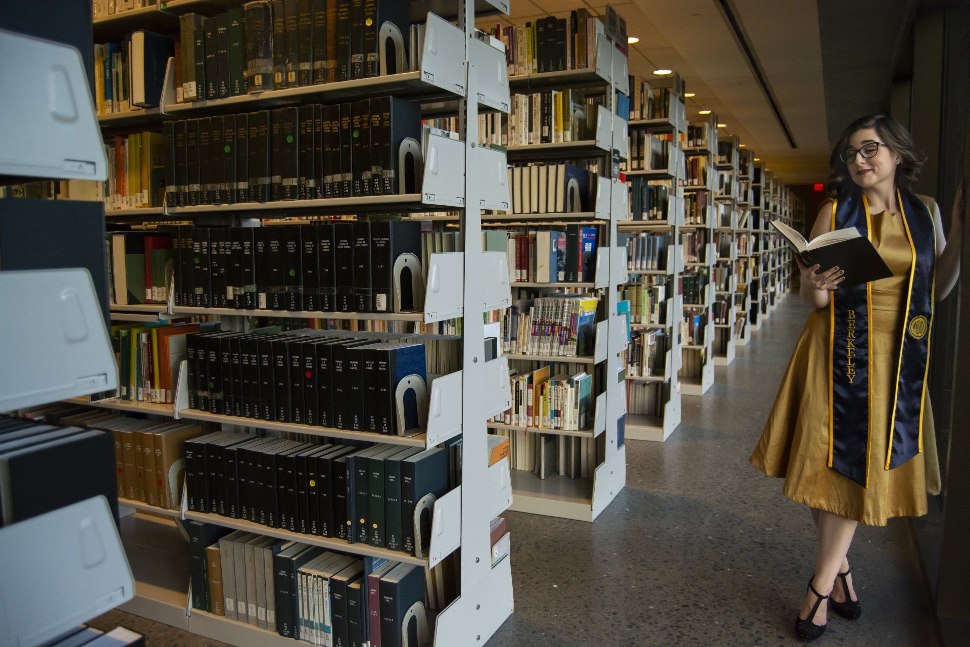 Woman in gold dress and sash reads a book in a library, leaning against a wall. Shelves of books fill the background.