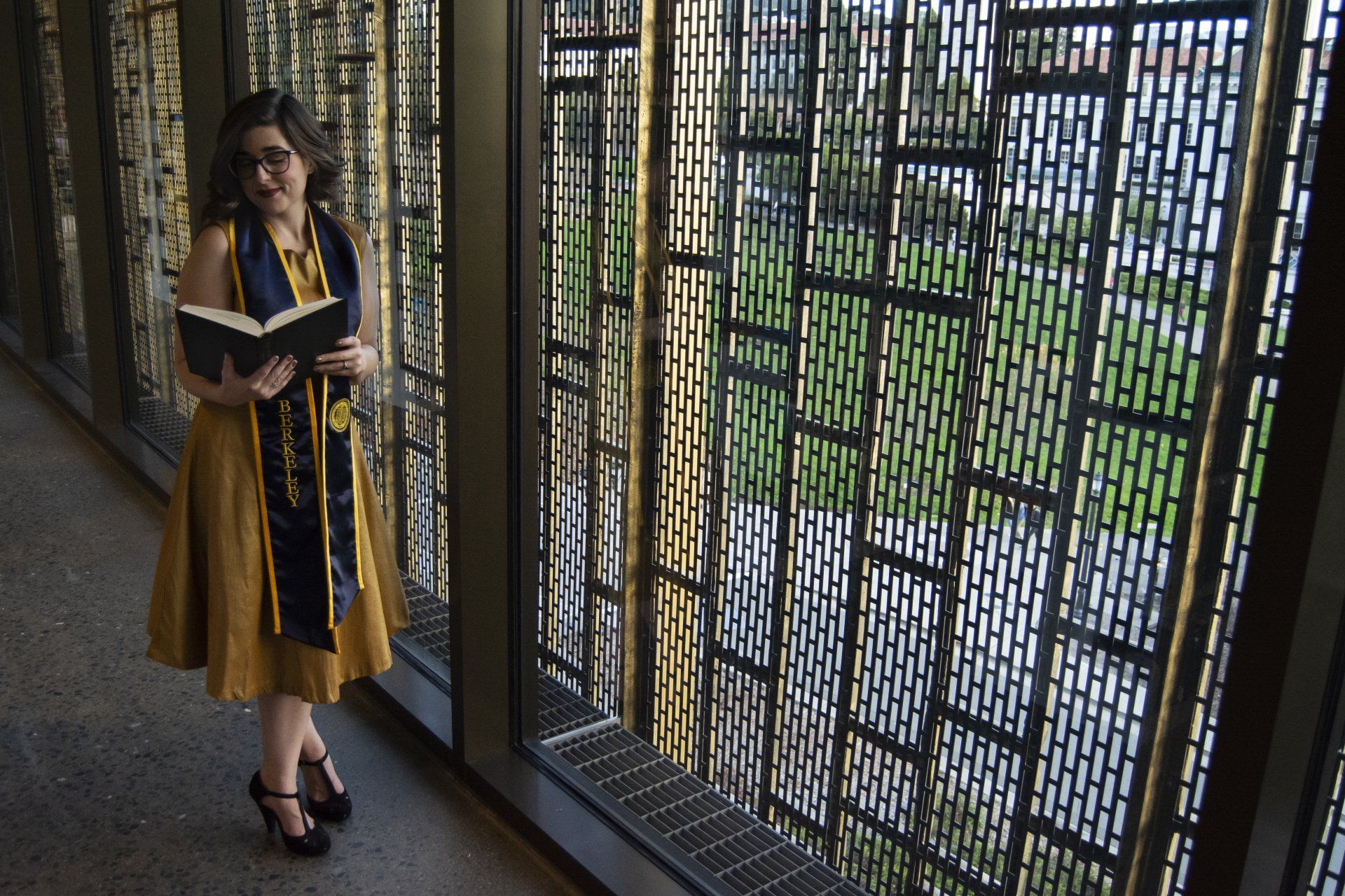 Woman in graduation attire holding a book, standing near a decorative metal screen.