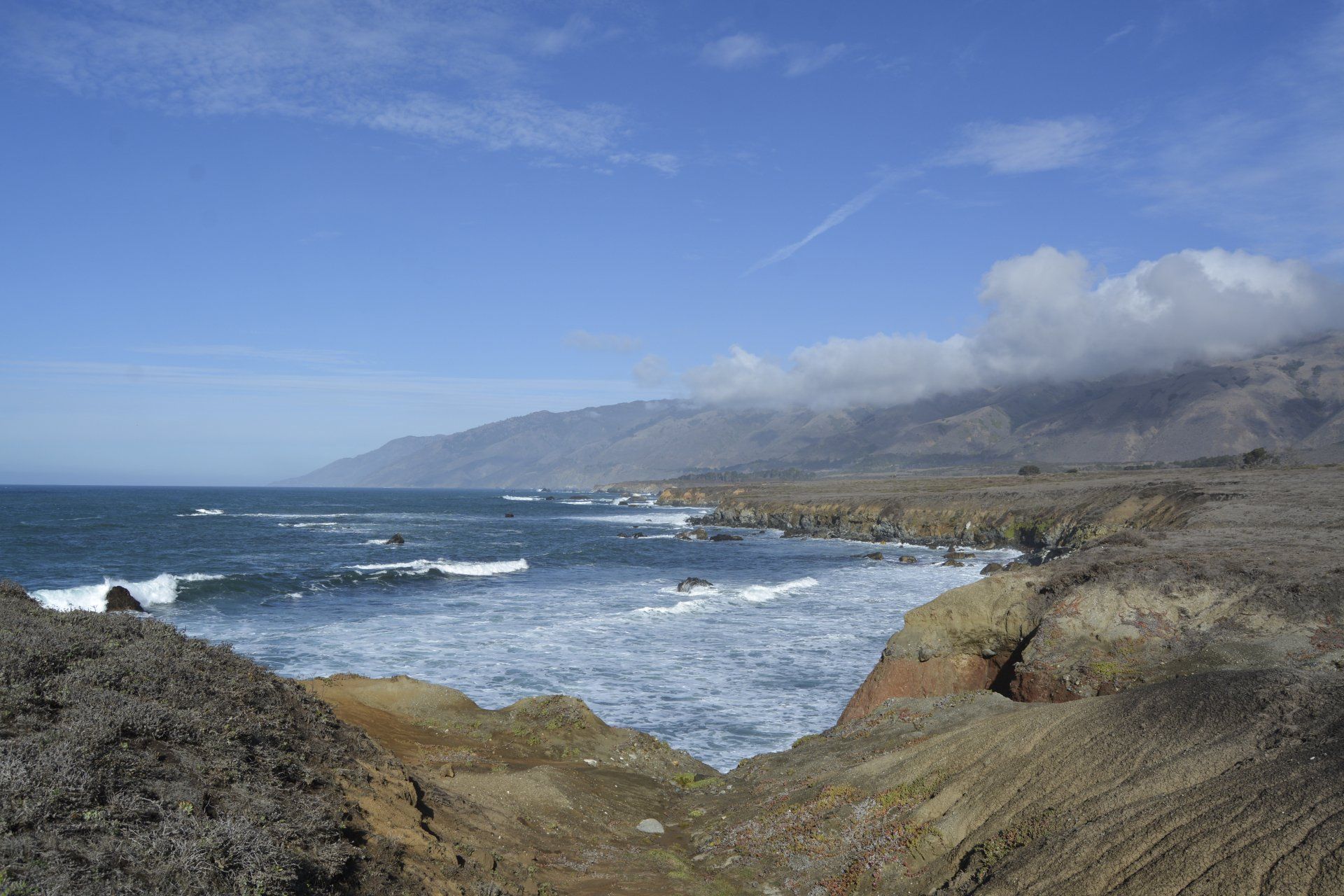 Rocky coastline with crashing waves under a partly cloudy blue sky.