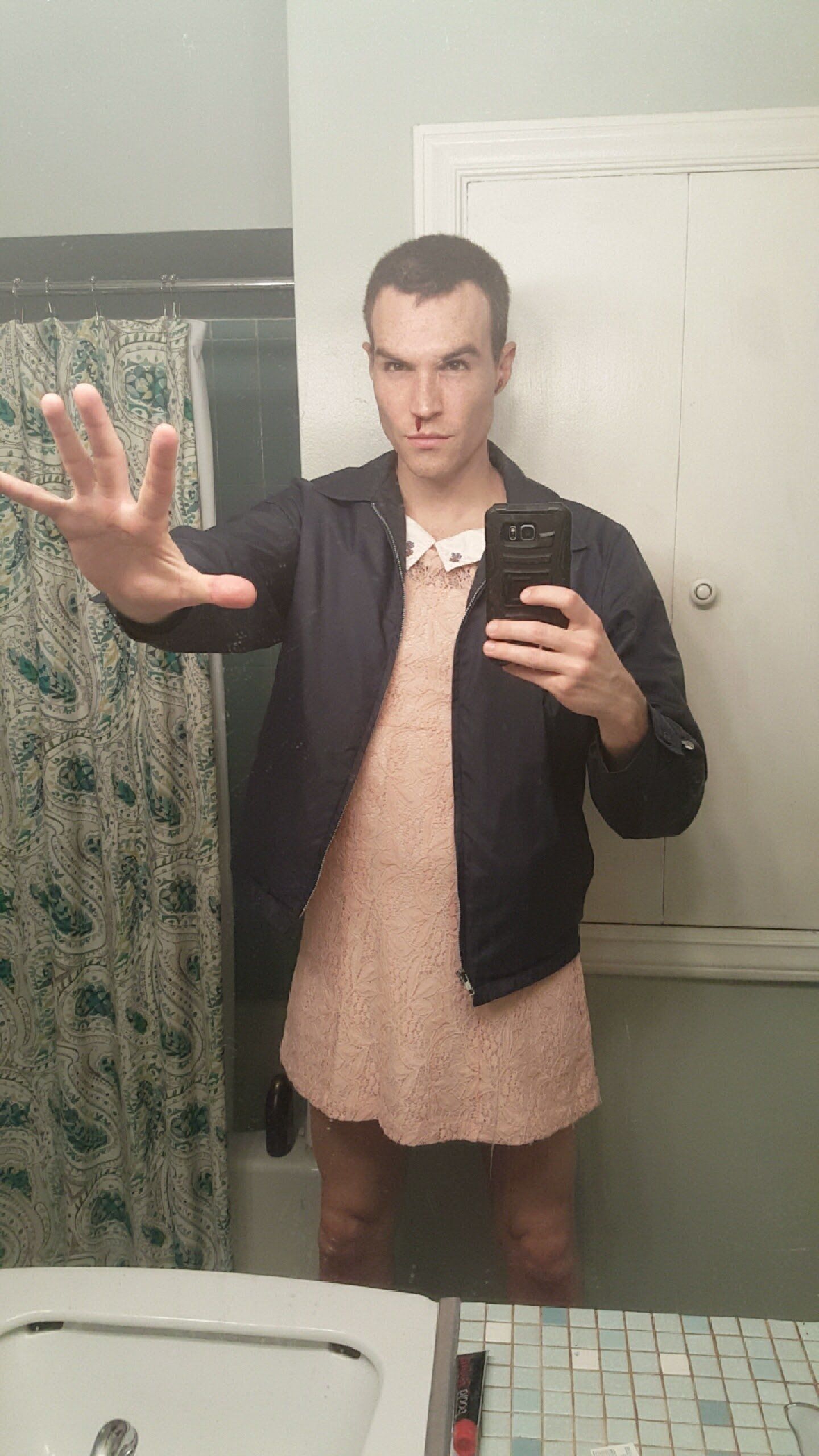 Person in a pink dress and navy jacket poses with hand outstretched, holding a phone, in a bathroom.